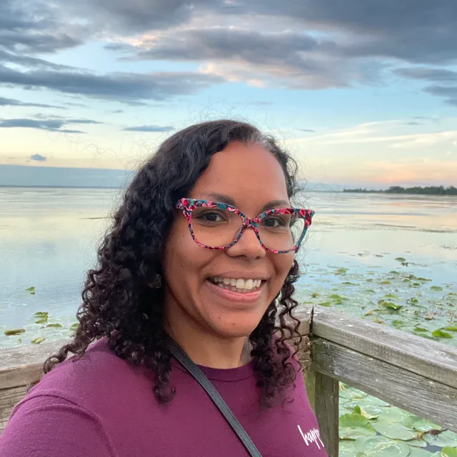 Travel Advisor Andrea Woodard with a red shirt in front of a lake at sunset.