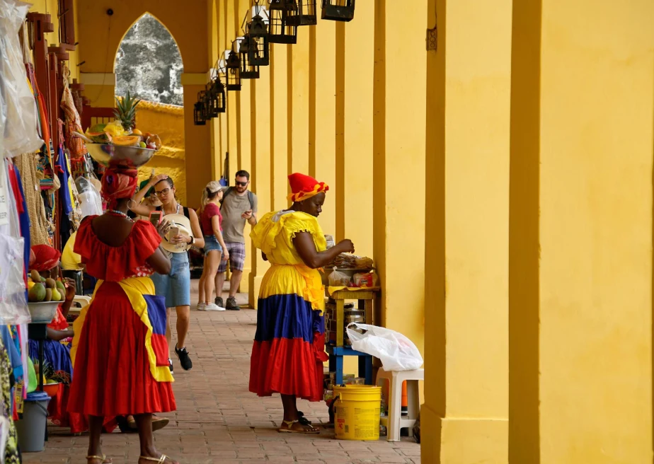 a street lined with yellow columns with various market stalls and women dressed in brightly colored dresses