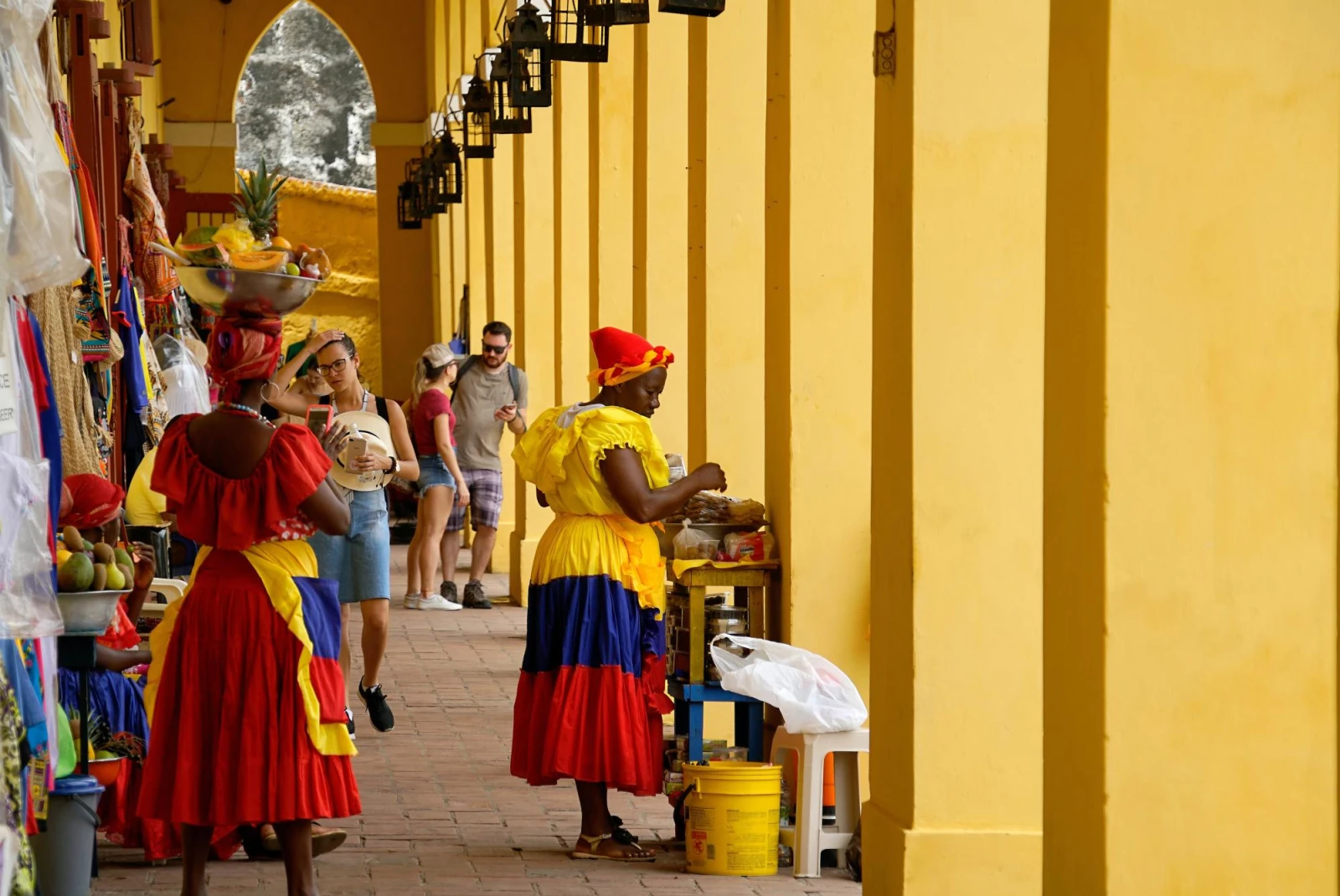 a street lined with yellow columns with various market stalls and women dressed in brightly colored dresses