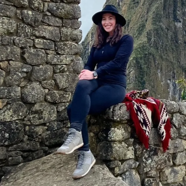Travel Advisor Anne Littman is wearing a black hat and sitting on a rocky wall Infront of a mountain.