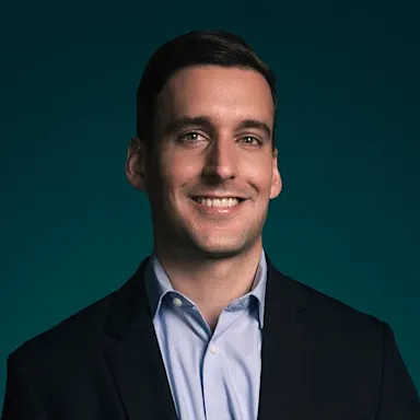 Professional headshot of a smiling man in a black suit jacket and light blue shirt against a teal background.