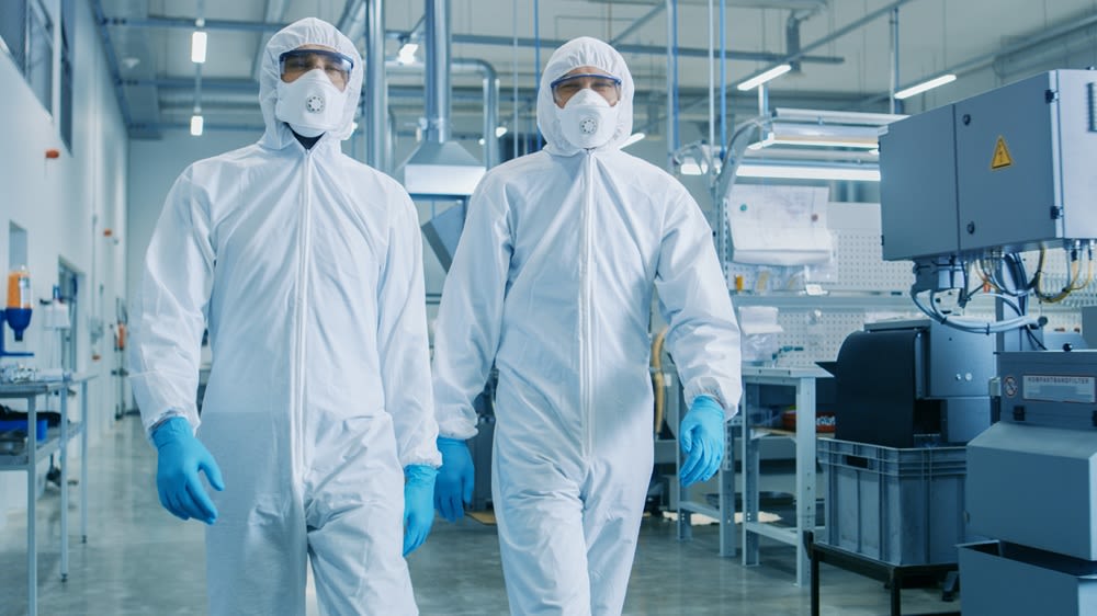 Two workers wearing hazmat clean room bunny suits, blue latex gloves, face masks, and goggles, walking through a clean room environment.
