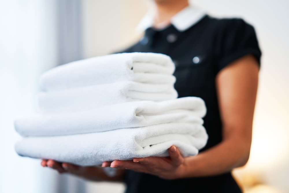 A blurred background of a hotel maid holding a stack of white bath towels in-focus in the foreground of the picture.