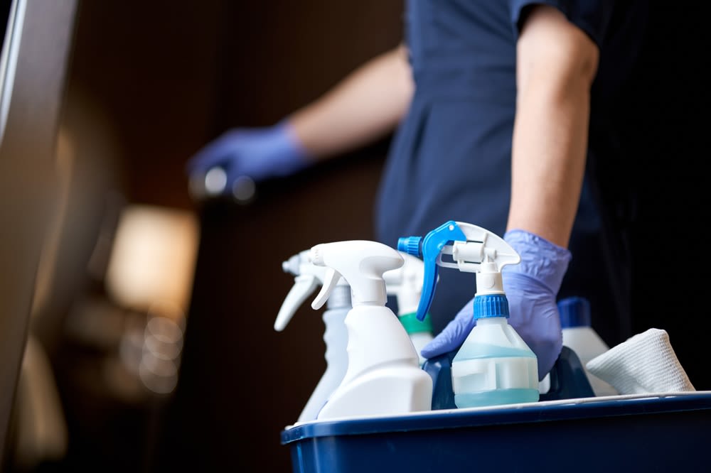 A cleaner entering a hotel room with a collection of cleaning sprays and towels.