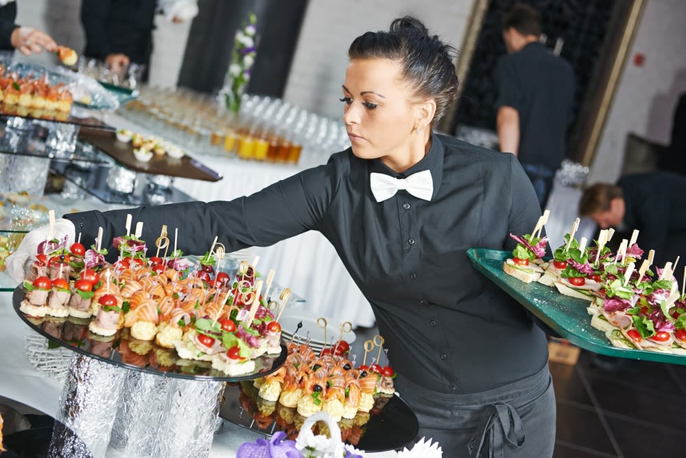 A white woman caterer dressed in a black outfit with white bowtie balances plates of food.