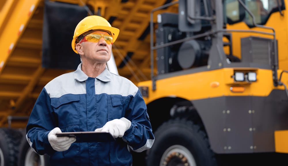 A man wearing a hard hat, goggles, and an industrial work jacket holding a tablet and standing in front of a dump truck.