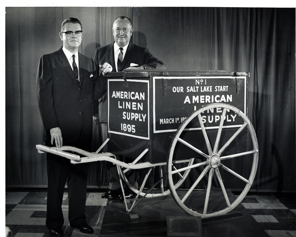 A black and white photo of two historical Alsco Uniforms executives standing next to an old company handcart.