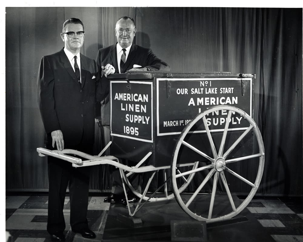 A black and white photo of two historical Alsco Uniforms executives standing next to an old company handcart.