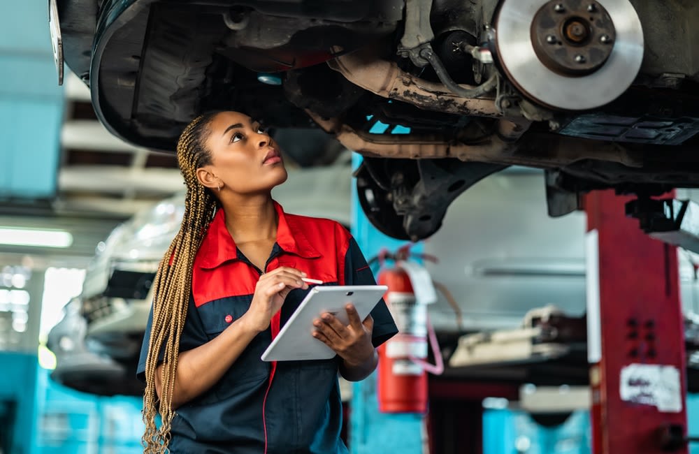 A young Black woman working as a mechanic examining a car that's lifted up. She is dressed in a mechanic's shop suit and is holding a tablet.