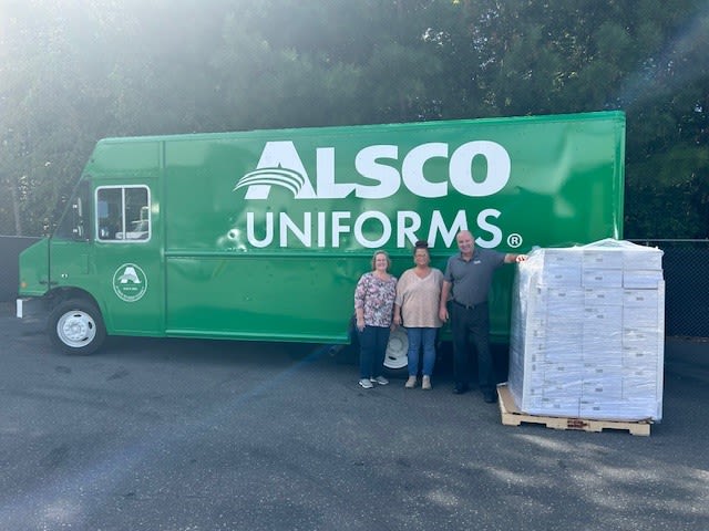 Alsco Uniforms employees standing next to a green delivery truck and a pallet of first aid kits.