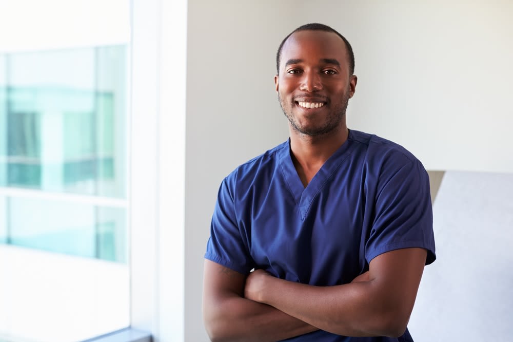 A young Black man dressed in purple scrubs smiling and standing with his arms folded.