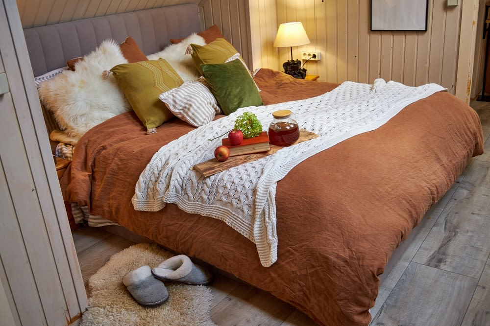 A cozy bed with lots of pillows, a plush comforter and quilt, and a tray with a book, fruit, and a jar of tea.