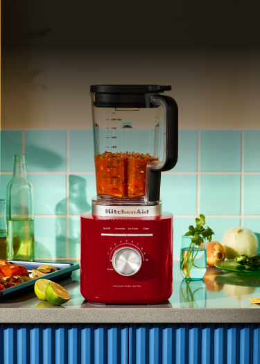 A red KitchenAid® blender next to a pan of roasted veggies and blended salsa inside