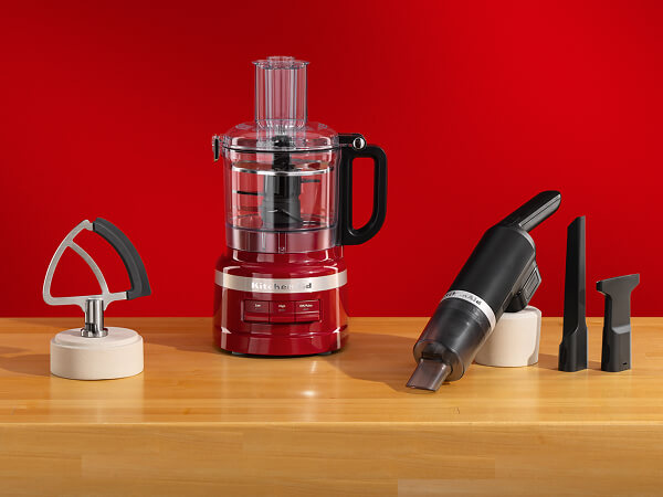 Flat edge beater, red food processor and a KitchenAid Go® Cordless countertop vacuum with different attachments on a wooden counter. 
