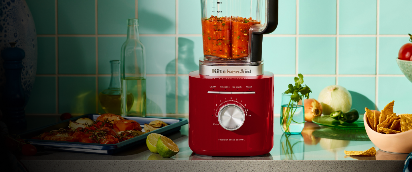 A red KitchenAid® blender next to a pan of roasted veggies and blended salsa inside