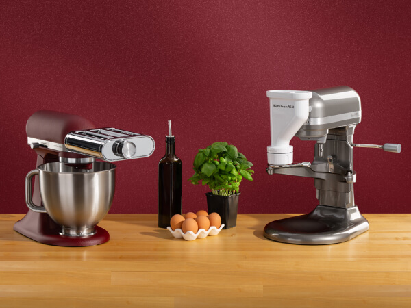 A red stand mixer with a pasta attachment and a silver stand mixer with a meat grinding attachment on a wooden counter with eggs, olive oil and basil