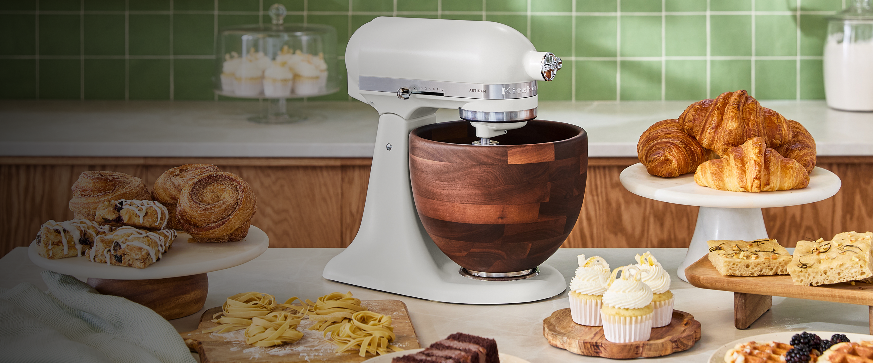 A Porcelain White KitchenAid® stand mixer with a wood bowl on the counter, near a slew of pastries.
