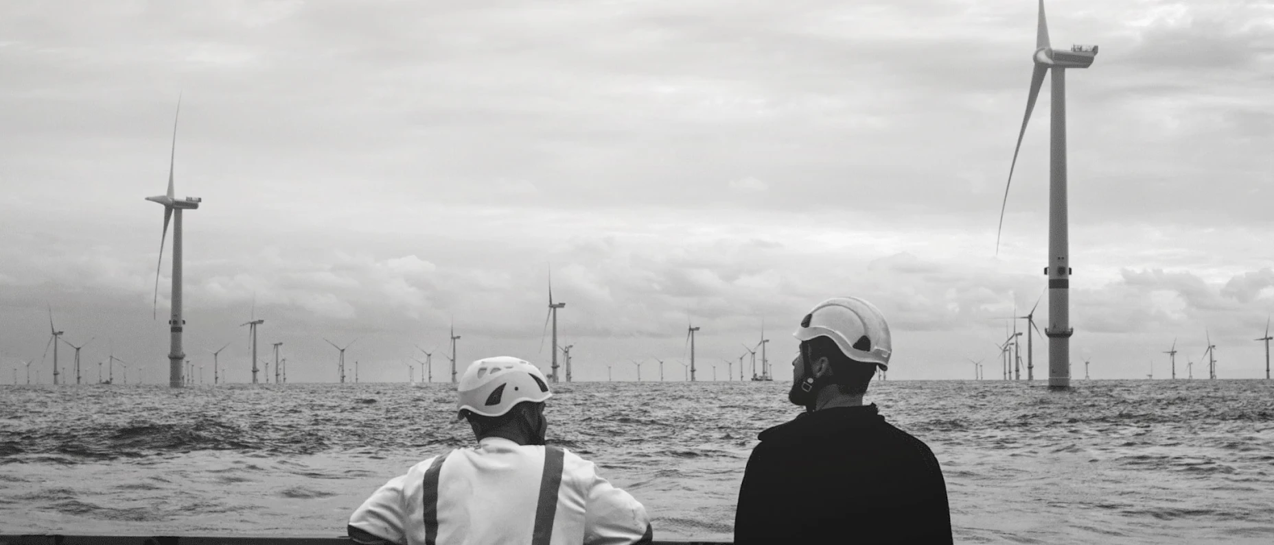 Schwarz-Weiß-Bild mit zwei Arbeitern, die auf einen Windpark im Wasser schauen.