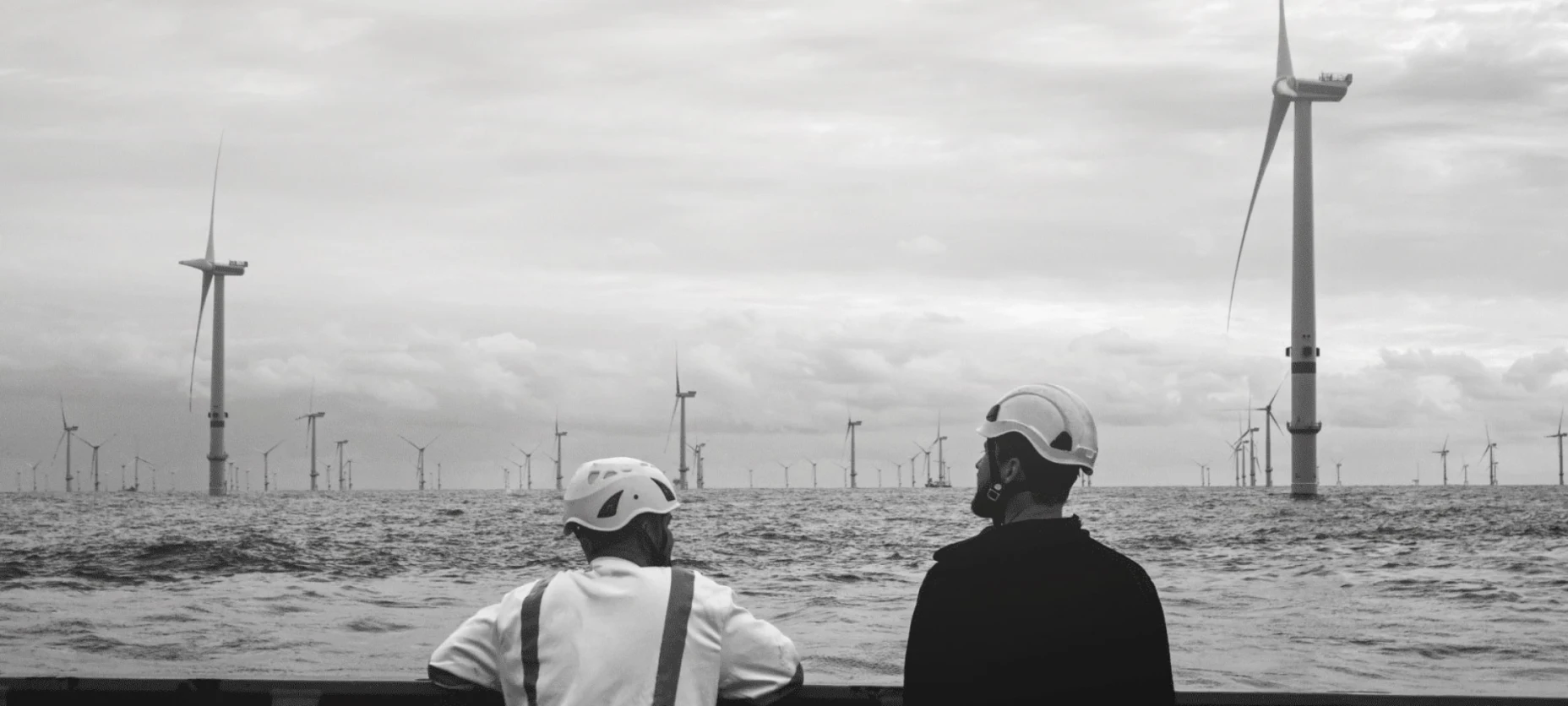 Schwarz-Weiß-Bild mit zwei Arbeitern, die auf einen Windpark im Wasser schauen.