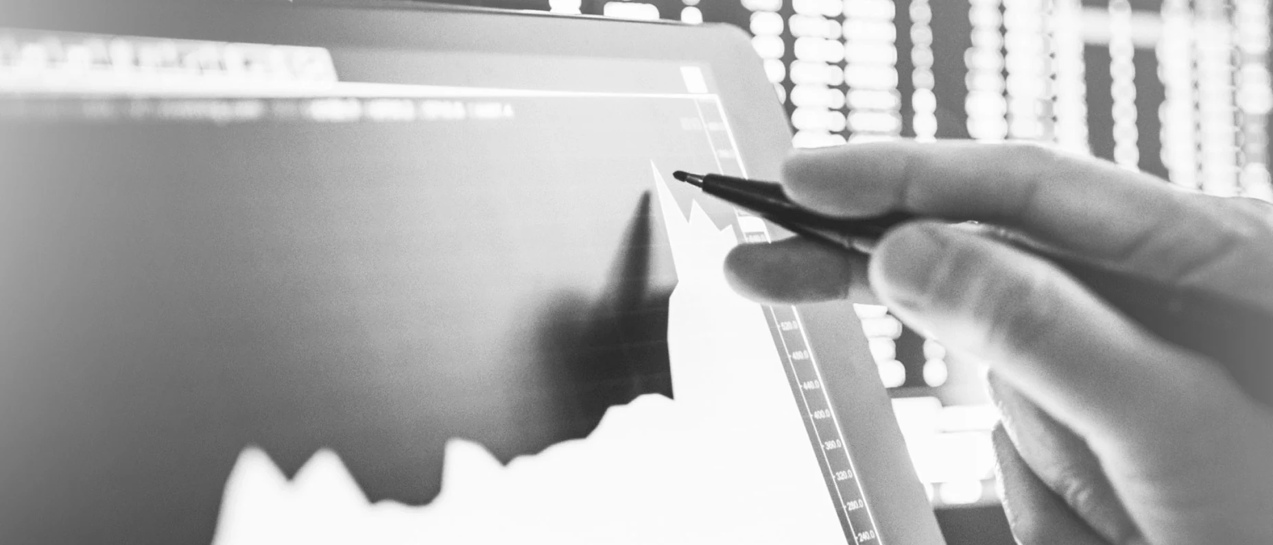 Schwarz-Weiß-Bild auf dem ein Kursverlauf auf einem Tablet zu sehen ist. Eine Hand mit einem Stift zeigt auf das Tablet.