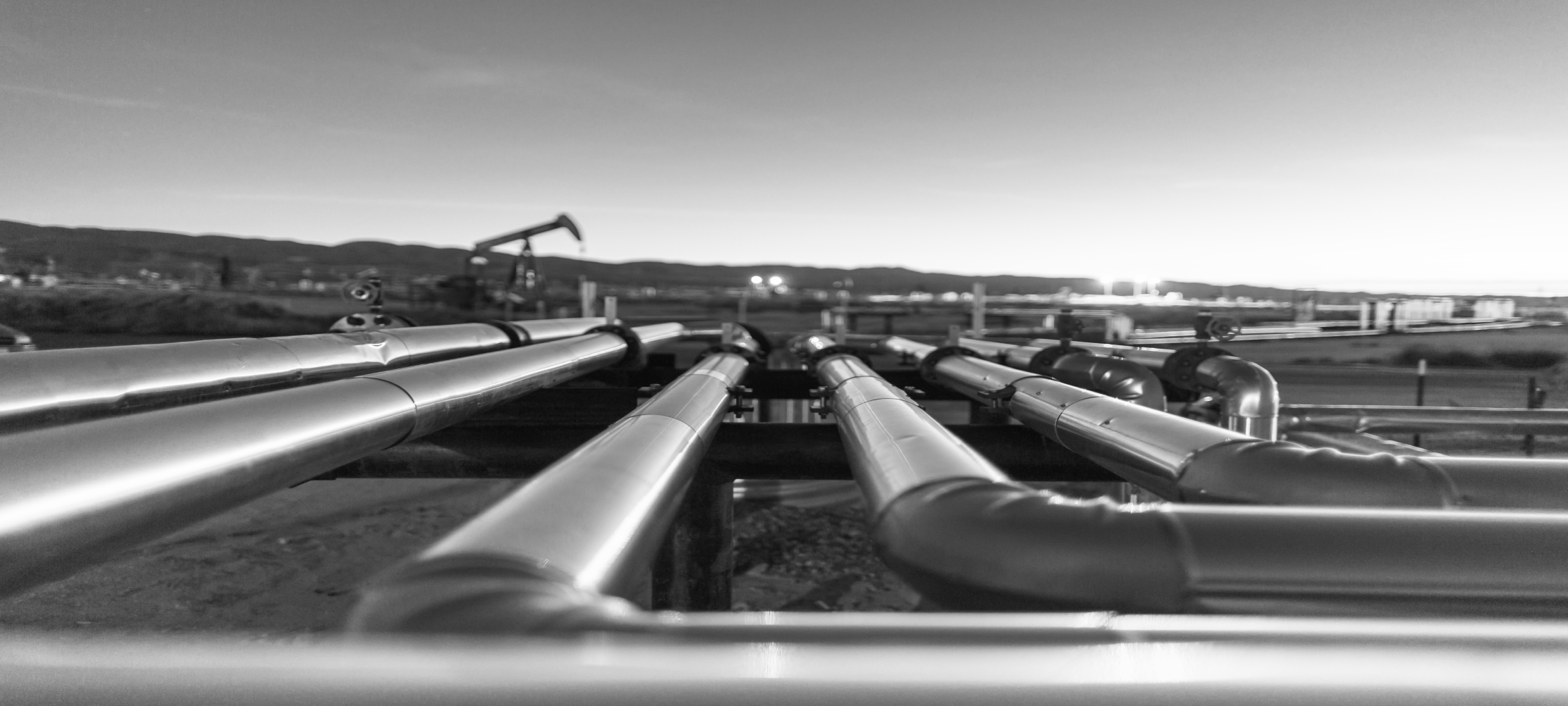 Schwarz-Weiß-Bild von einem Rohleitungsnetz im Vordergrund. Im Hintergrund sind ein Ölbohrer und eine hügelige Landschaft zu sehen.