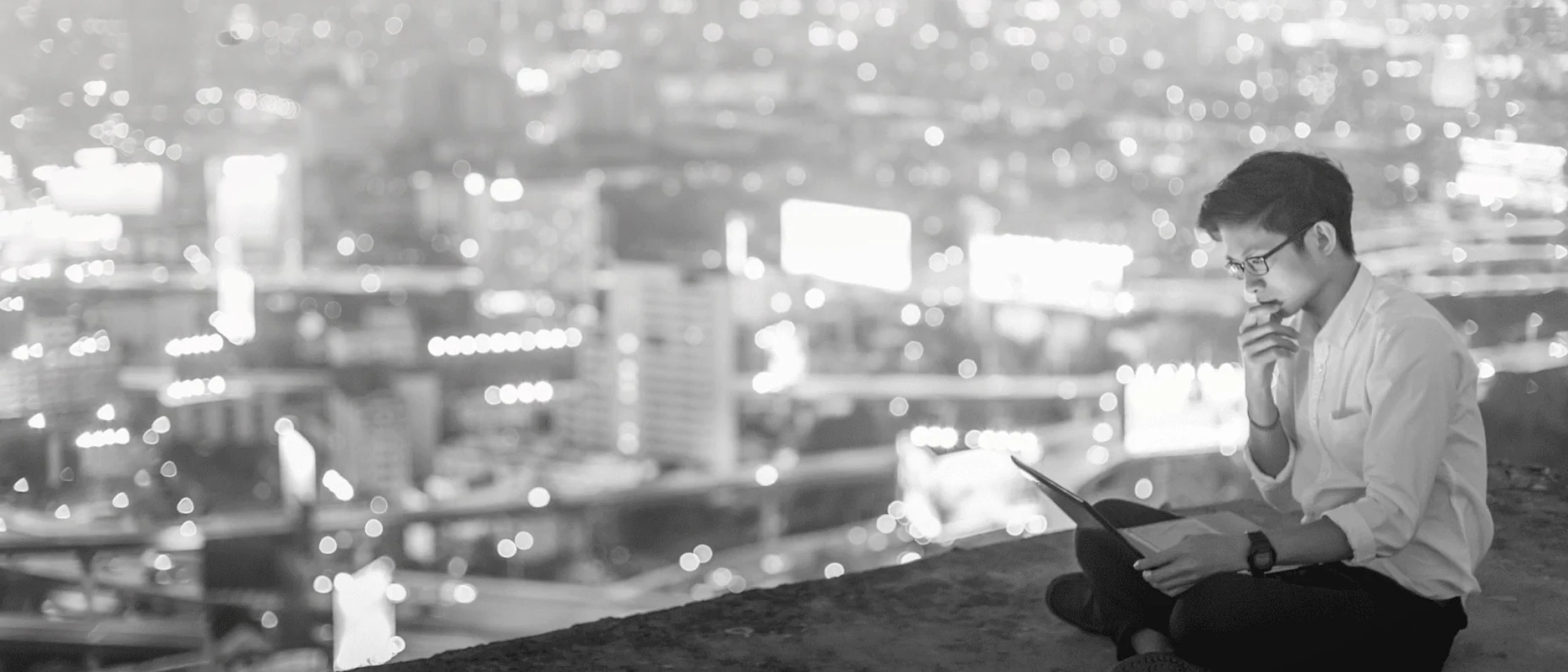 Schwarz-Weiß-Bild mit einem Mann sitzend und Laptop in der Hand über den Dächern und einem Ausblick auf die Skyline einer Stadt im Hintergrund.