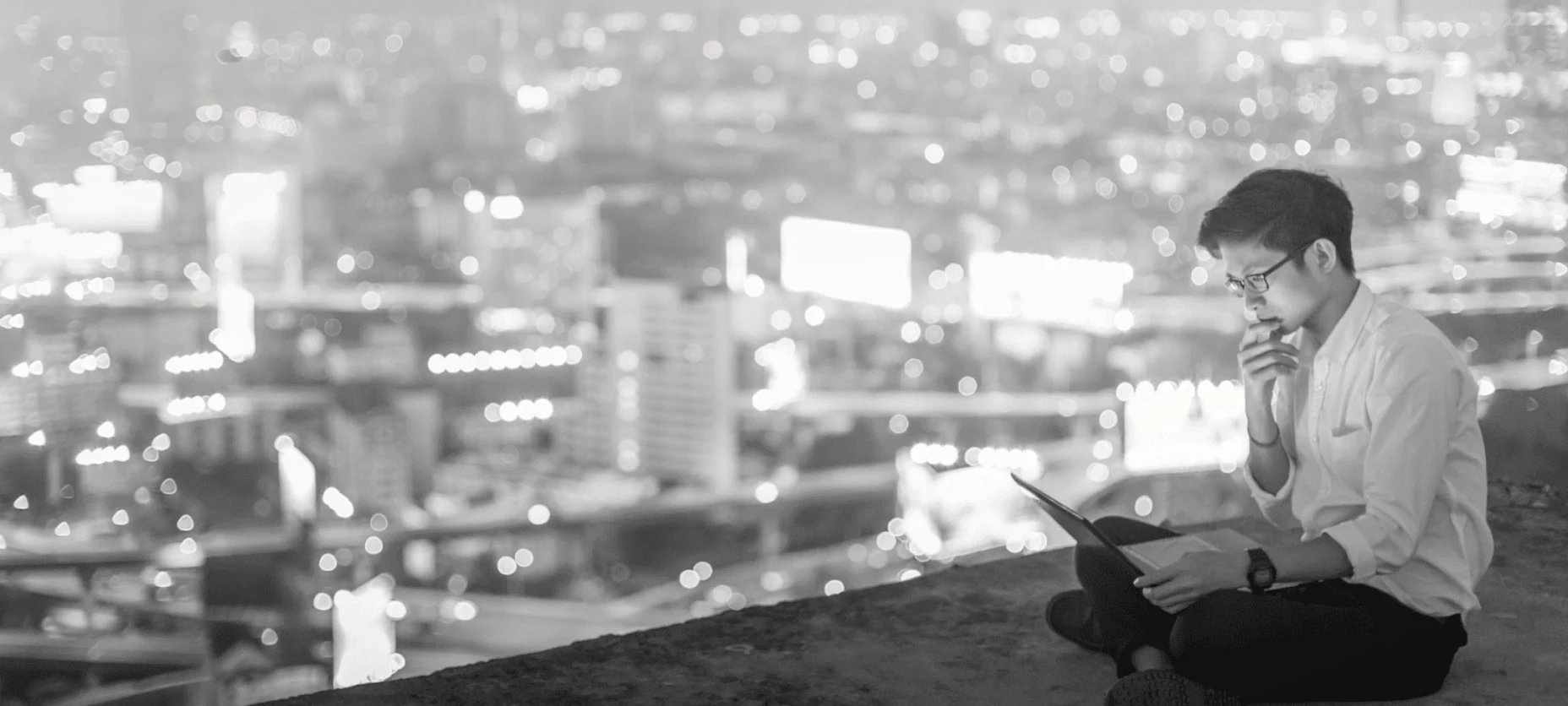 Schwarz-Weiß-Bild mit einem Mann sitzend und Laptop in der Hand über den Dächern und einem Ausblick auf die Skyline einer Stadt im Hintergrund.