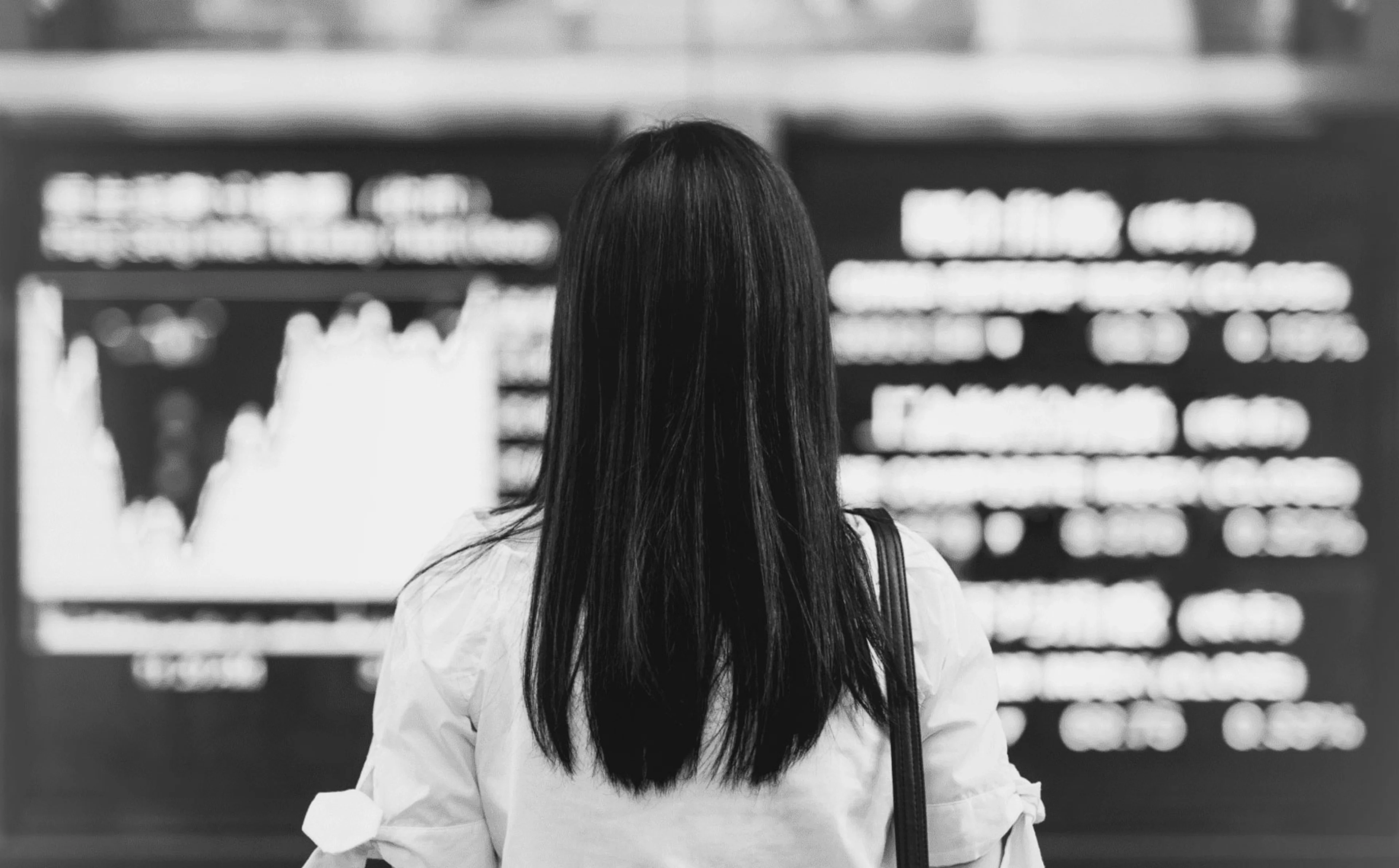 Frau steht vor einer Displaywand, welche die aktuelle Börsenkurse zeigt