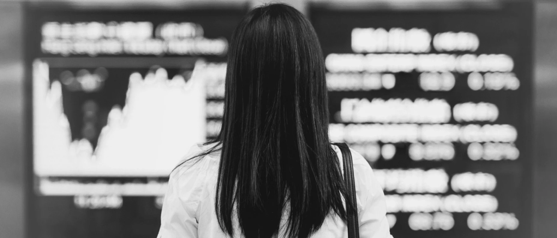 Schwarz-Weiß-Bild einer Frau, welche vor einer Displaywand steht und sich die aktuellen Börsenkurse anschaut.