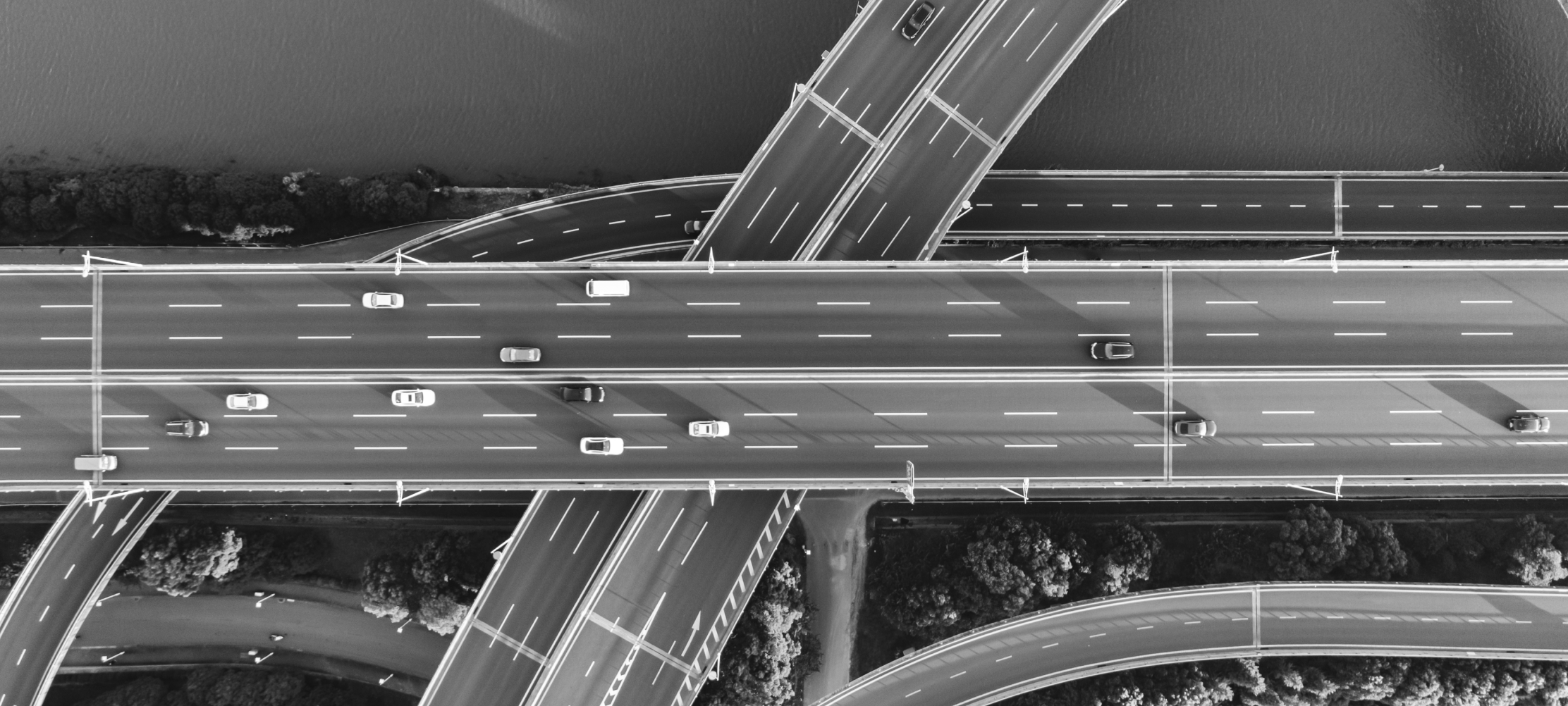 Schwarz-Weiß-Luftaufnahme einer mehrstöckigen Straßenkreuzung mit mehreren Fahrbahnen, Autos und geschwungenen Auffahrten über einem bewaldeten Gelände und einem Gewässer.