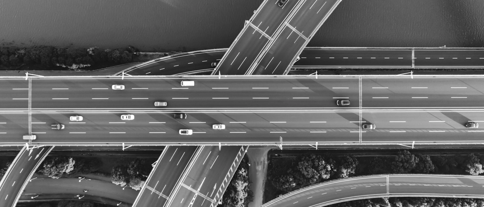 Schwarz-Weiß-Luftaufnahme einer mehrstöckigen Straßenkreuzung mit mehreren Fahrbahnen, Autos und geschwungenen Auffahrten über einem bewaldeten Gelände und einem Gewässer.