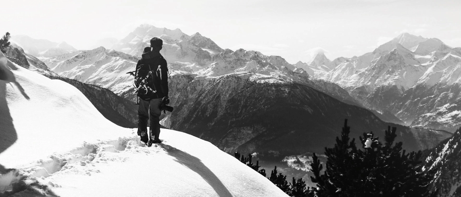 Schwarz-Weiß-Bild mit einem Bergsteiger, der auf einem schneebedeckten Hang steht und in die Ferne guckt. Im Hintergrund ein Ausblick auf die Berge.