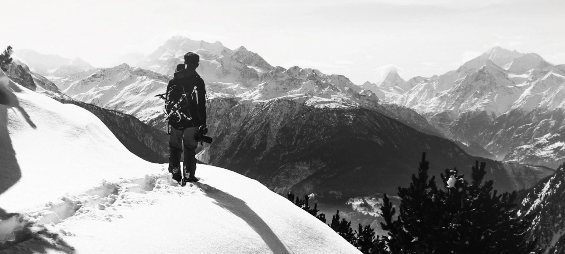Schwarz-Weiß-Bild mit einem Bergsteiger, der auf einem schneebedeckten Hang steht und in die Ferne guckt. Im Hintergrund ein Ausblick auf die Berge.