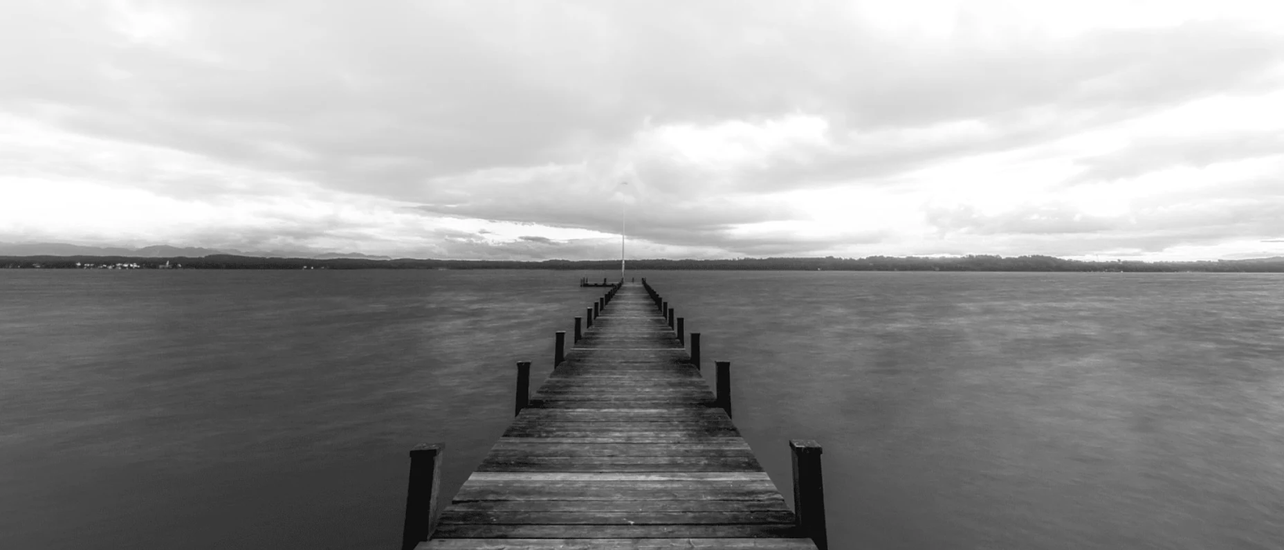 Schwarz-Weiß-Bild mit einem Steg an einem See und bewölktem Wetter.