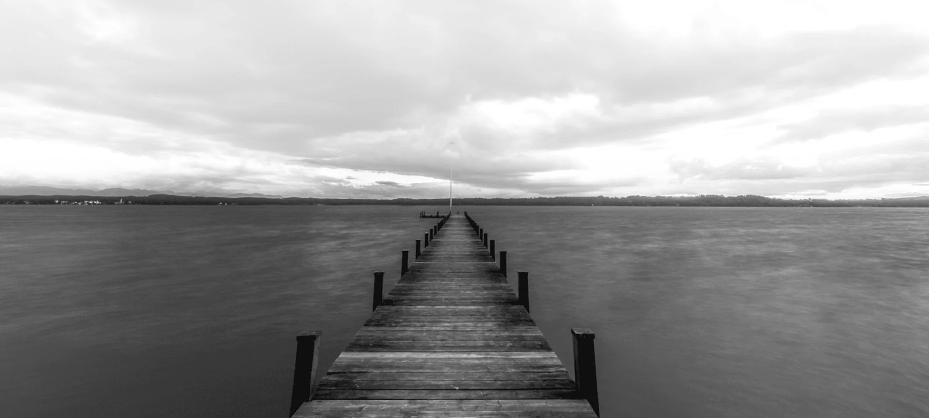Schwarz-Weiß-Bild mit einem Steg an einem See und bewölktem Wetter.