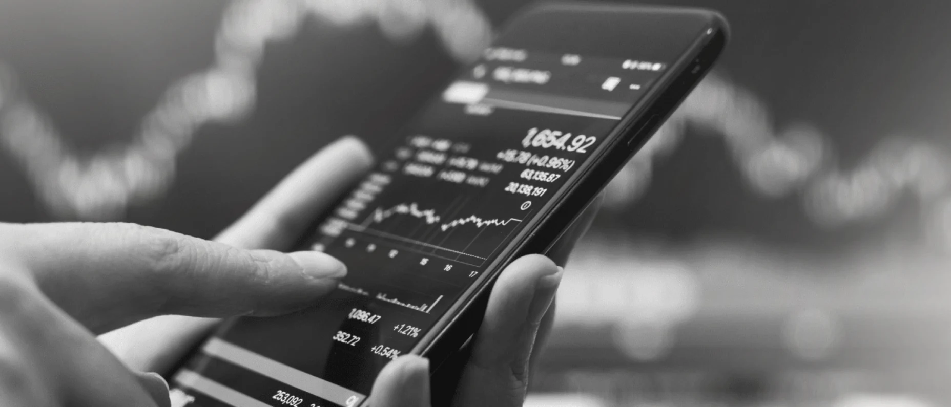Schwarz-Weiß-Bild einer Hand, die ein Smartphone hält auf dem ein Kursverlauf angezeigt wird.