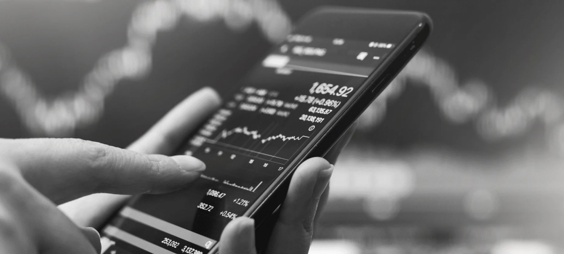 Schwarz-Weiß-Bild einer Hand, die ein Smartphone hält auf dem ein Kursverlauf angezeigt wird.
