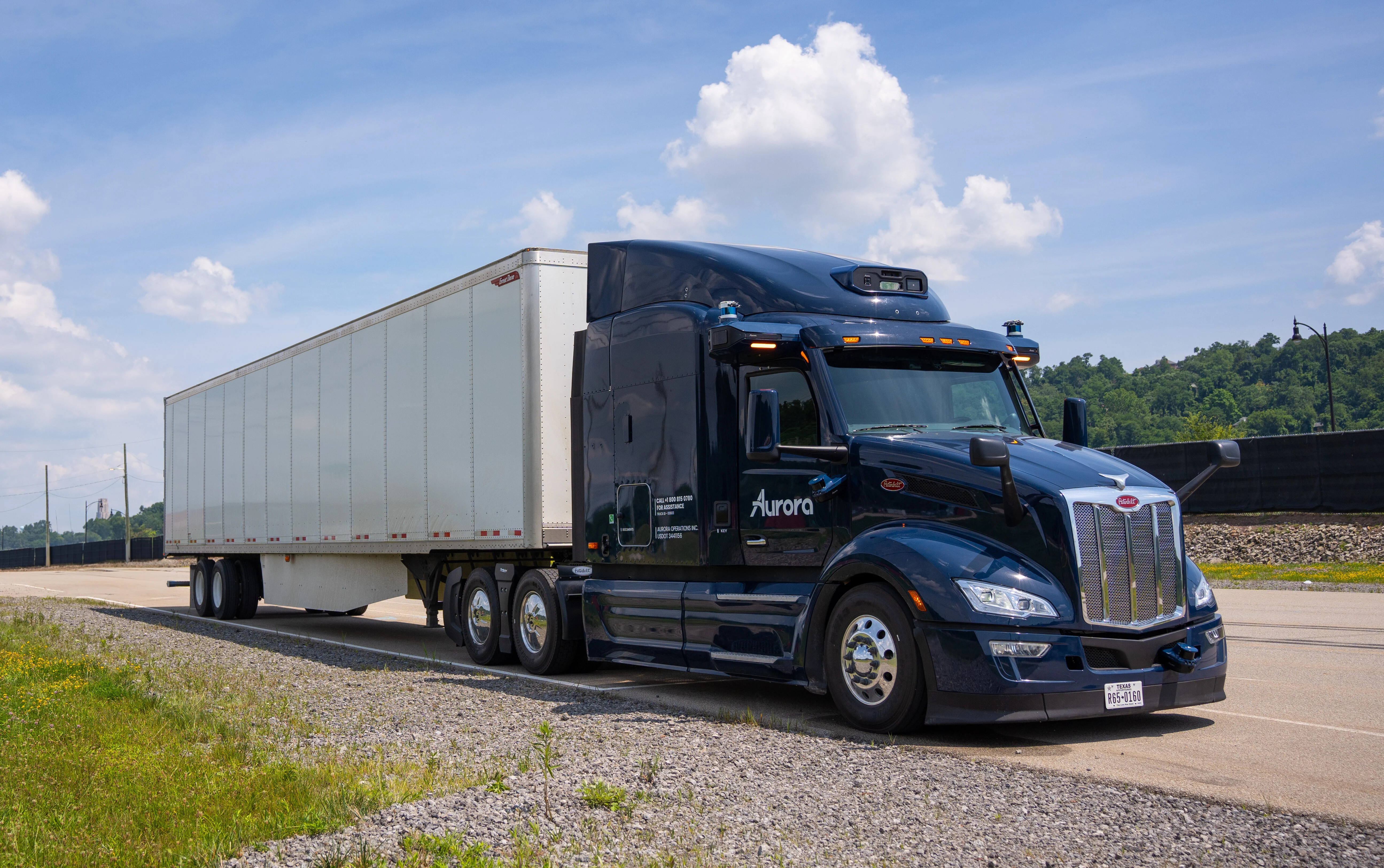 A dark blue Peterbilt semi-truck with the Aurora logo is driving on a highway under a partly cloudy sky. The truck is hauling a white trailer and is captured in motion on the asphalt road, surrounded by a grassy shoulder and distant trees. This image represents Aurora's technology in autonomous long-haul trucking