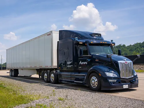 A dark blue Peterbilt semi-truck with the Aurora logo is driving on a highway under a partly cloudy sky. The truck is hauling a white trailer and is captured in motion on the asphalt road, surrounded by a grassy shoulder and distant trees. This image represents Aurora's technology in autonomous long-haul trucking