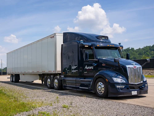 A dark blue Peterbilt semi-truck with the Aurora logo is driving on a highway under a partly cloudy sky. The truck is hauling a white trailer and is captured in motion on the asphalt road, surrounded by a grassy shoulder and distant trees. This image represents Aurora's technology in autonomous long-haul trucking