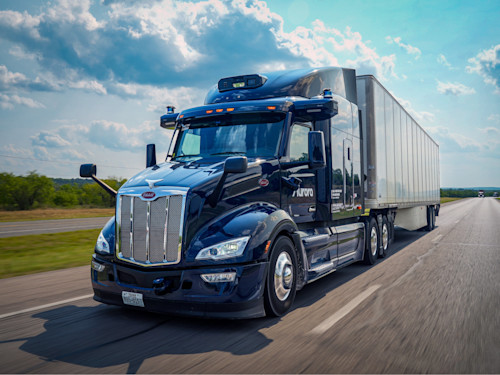 A sleek, dark blue Peterbilt semi-truck equipped with Aurora's autonomous driving technology travels on a multi-lane highway under a partly cloudy sky. The "Aurora" logo is visible on the side of the truck, signifying its self-driving capabilities and its role in the future of freight transportation.