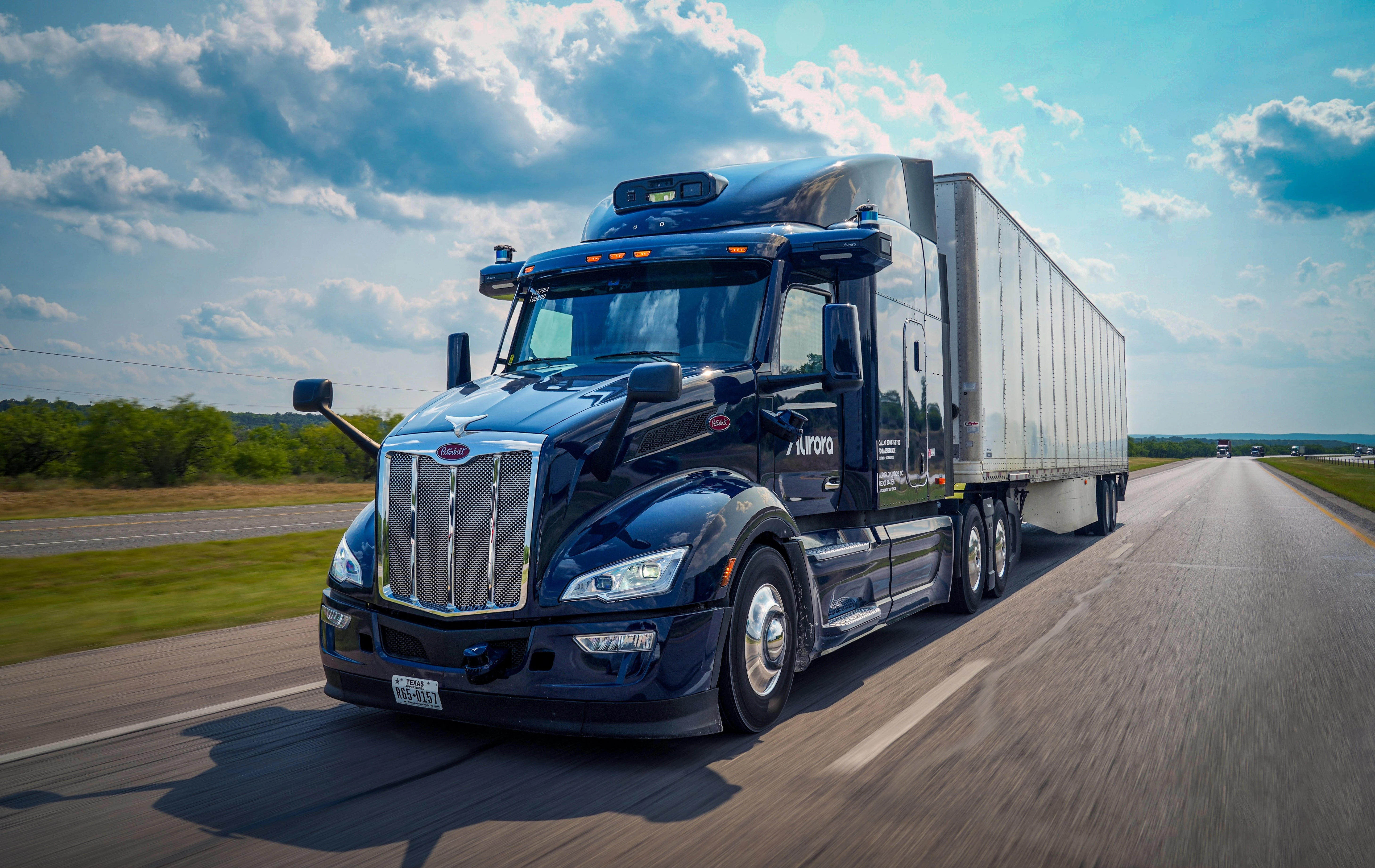 A sleek, dark blue Peterbilt semi-truck equipped with Aurora's autonomous driving technology travels on a multi-lane highway under a partly cloudy sky. The "Aurora" logo is visible on the side of the truck, signifying its self-driving capabilities and its role in the future of freight transportation.