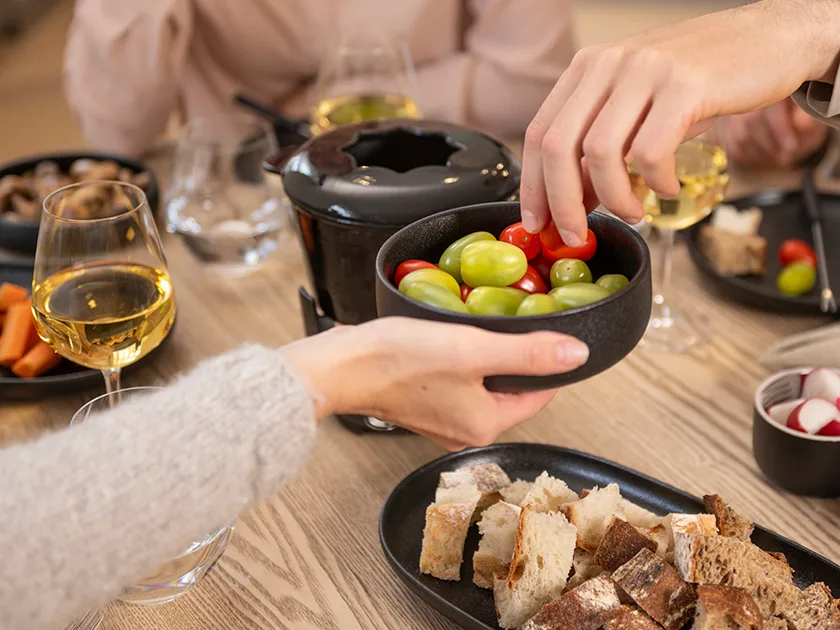 Naisen käsi ojentaa pöydän yli  kulhoa, jossa on miniluumutomaatteja. Toinen käsi ottaa tomaattia. Pöydän ympärillä on kaksi muutakin ihmistä, pöydällä on fonduepata, juomalaseja ja astioissa leipäkuutioita, porkkanatikkuja ja retiisejä