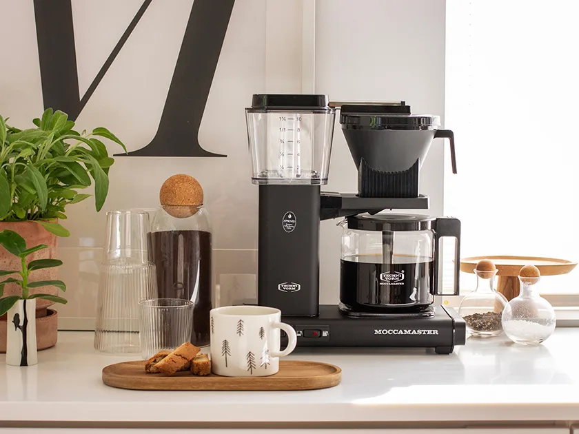 Keittiötasolla musta Moccamaster-kahvinkeitin, kahvikuppi, keksejä, lasiastioita ja yrtti saviruukussa