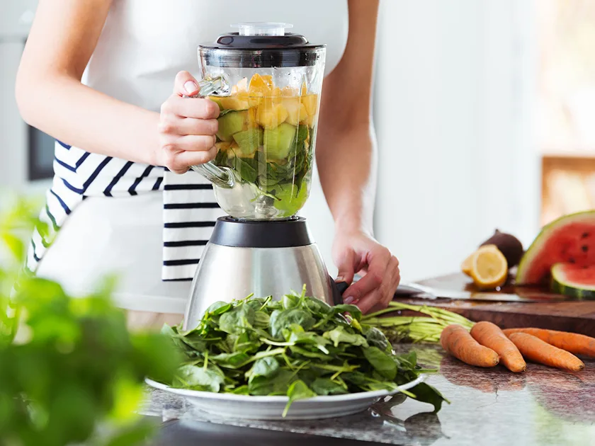 Nainen käyttää blenderiä, jossa on kasviksia ja hedelmää