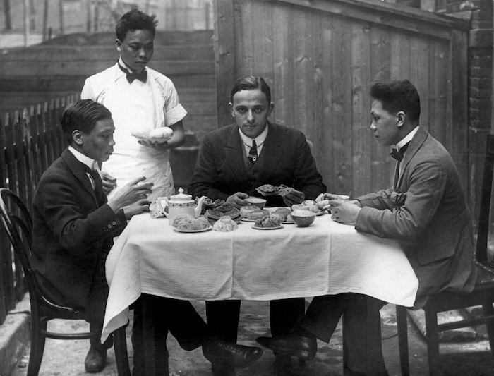 Chinezen in Rotterdam. Een fotoverslaggever van het tijdschrift Het Leven (midden) eet mee (lunch) met de Chinese restauranthouder Tsjang Kam Soi (links) in de Rotterdamse China-town in Katendrecht. Rotterdam, 1922
