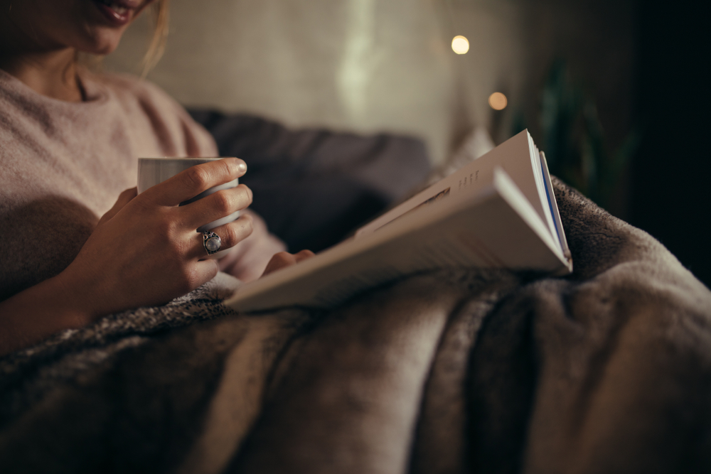 A woman is sitting on a bed in dim light holding a cup of tea and reading.