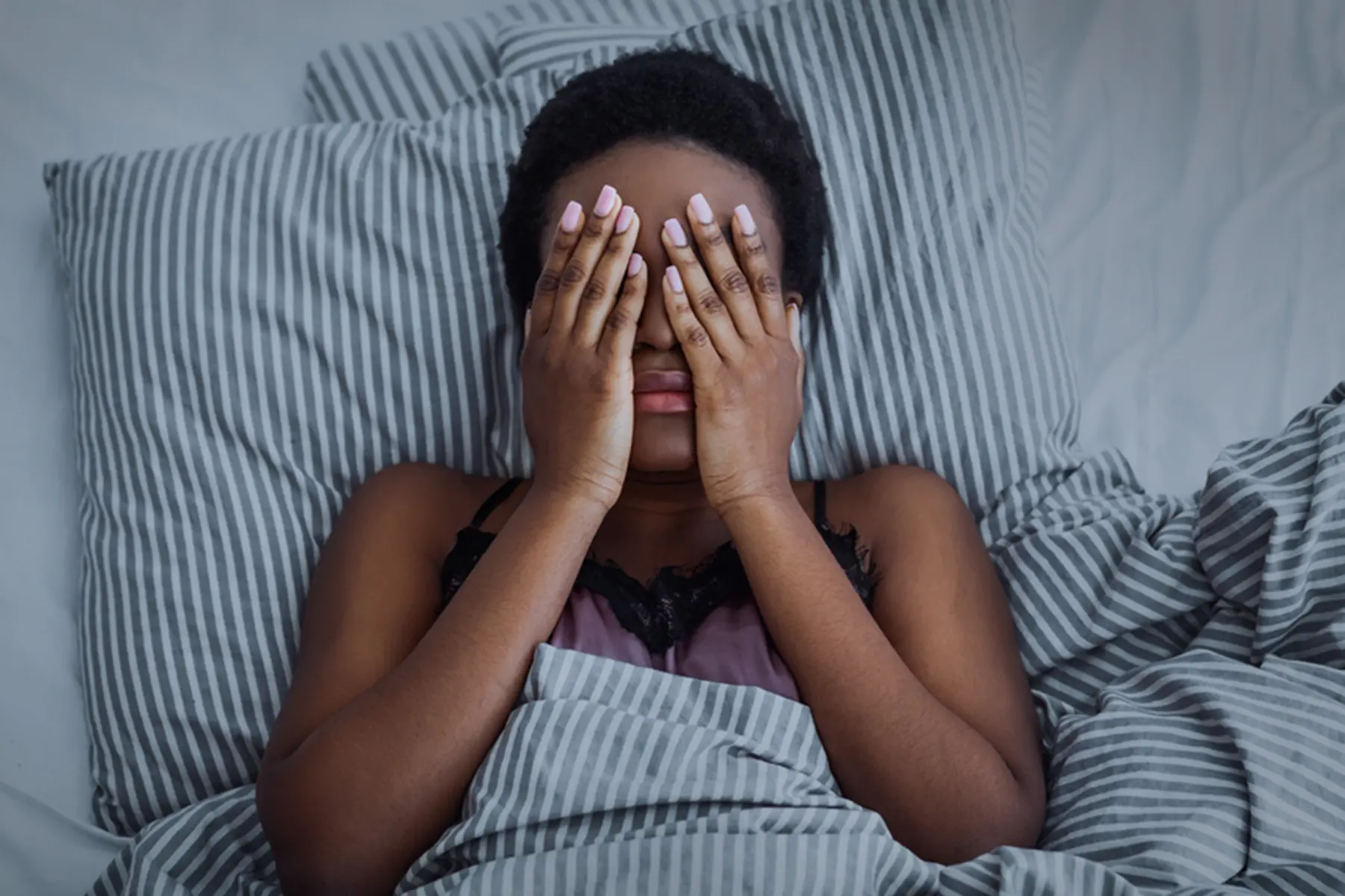   A woman lies in bed on blue and white striped sheets. Her hands are over her eyes in exhaustion, frustration, sadness, fear, or anxiety.