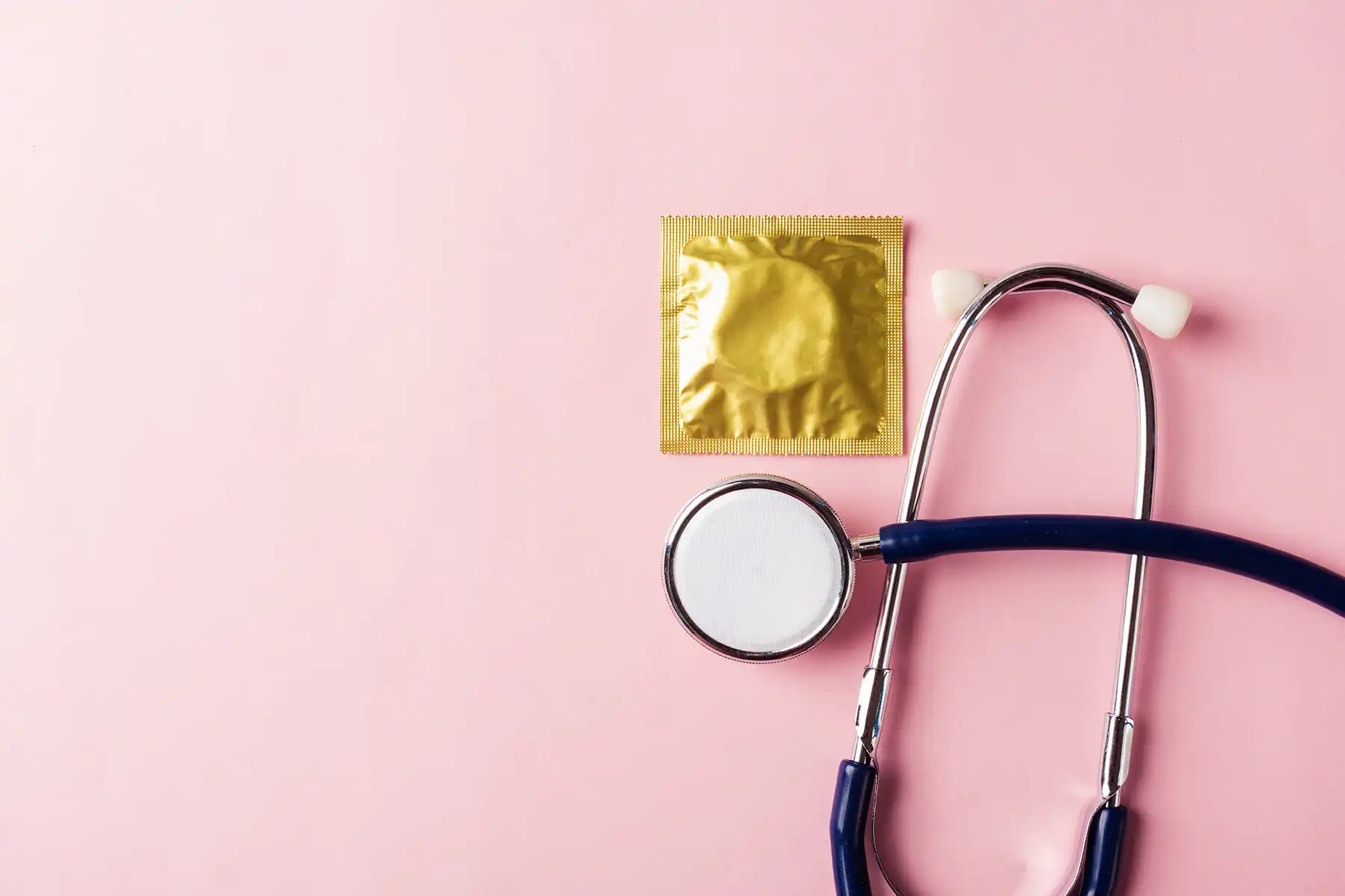 A condom with a gold wrapper and a stethoscope on a pink background.