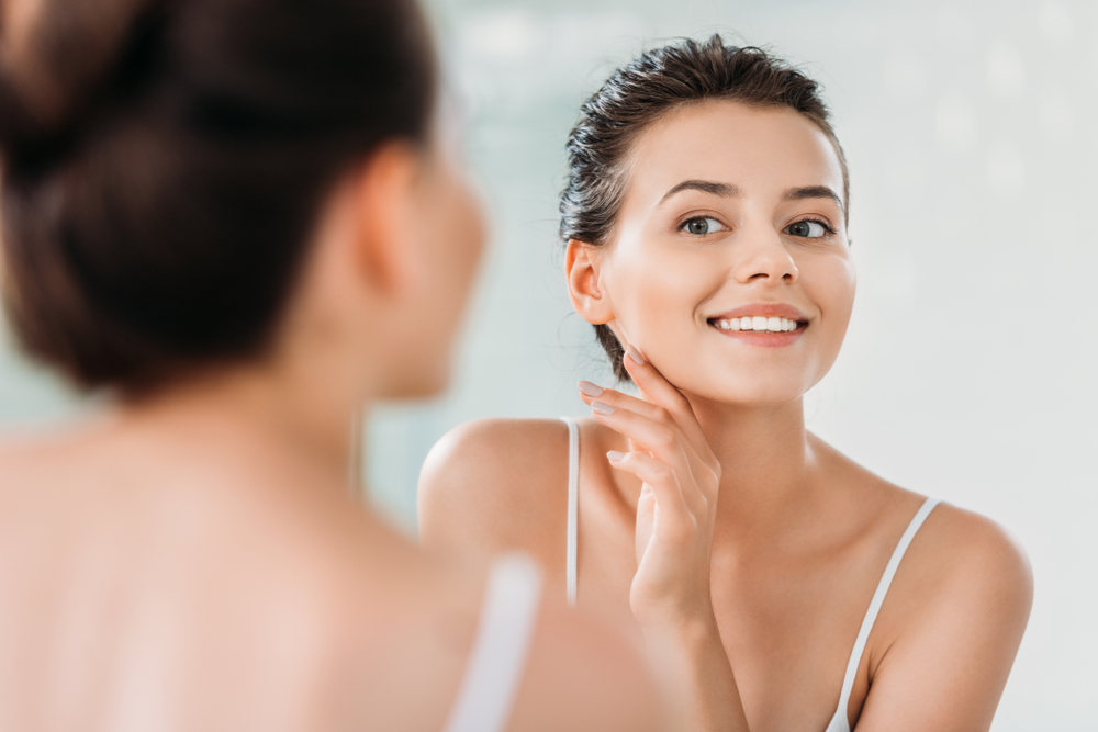 A young woman with dark hair pulled back is admiring her skin in the mirror. 